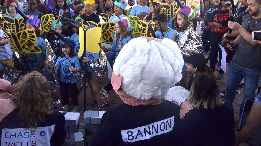 Children with paper butterflies and bird hats face effigies of Trump Administration officials.